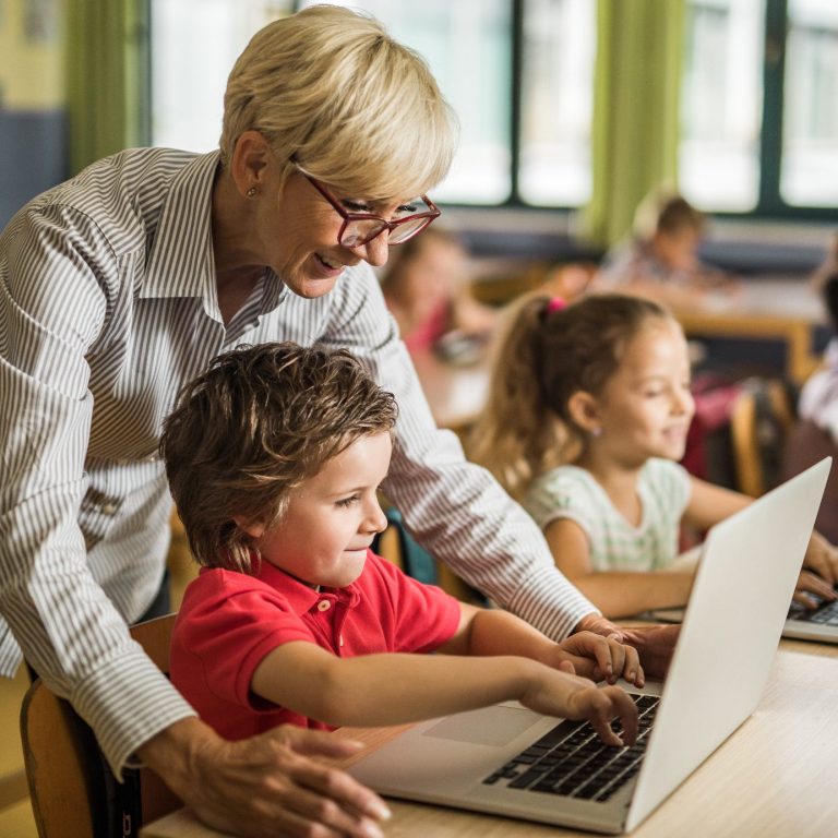 Lehrerin unterstützt Schülerinnen und Schüler im Umgang mit Computern in einer Klasse.