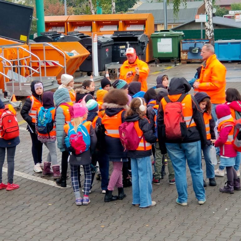 Eine Gruppe von Kindern besucht den Recyclinghof in Wilmersdorf-Charlottenburg. Die Kinder tragen alle orangefarbene  Westen. So sind sie gut sichtbar. Sie lauschen den spannenden Ausführungen zweier Mitarbeiter zum Thema Recycling. Die Kinder stehen vor riesengroßen Sammelbehältern.